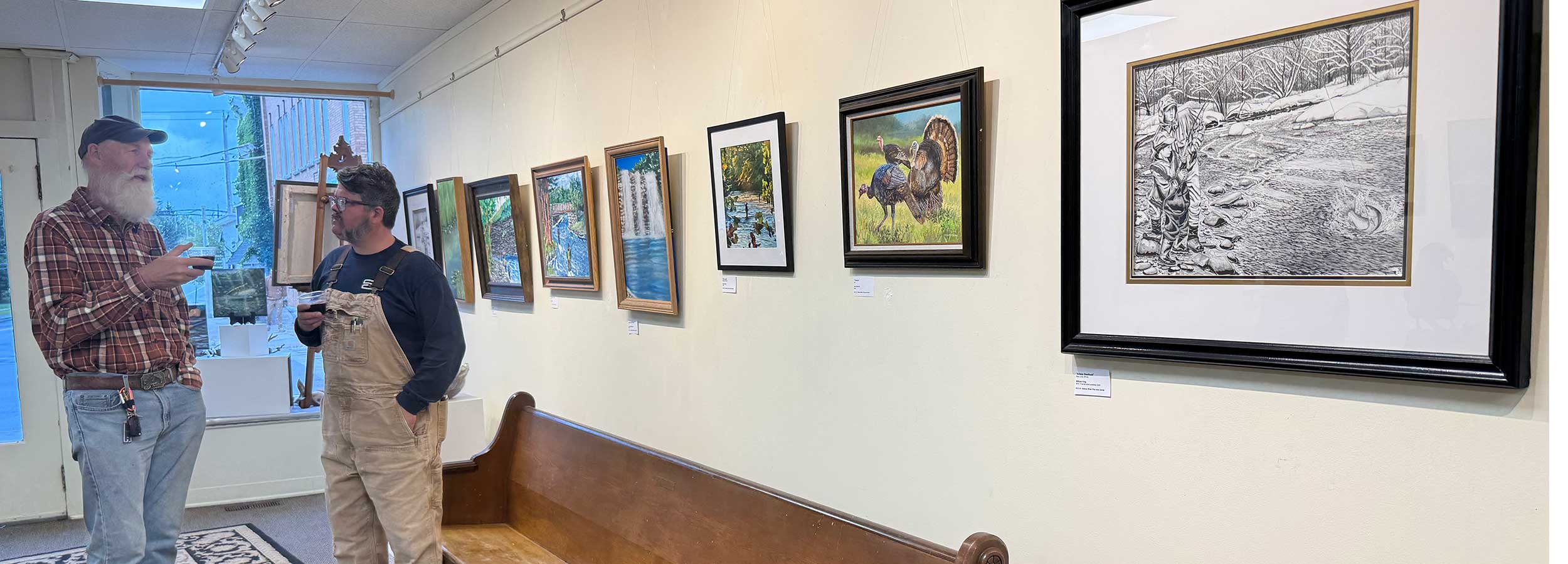 a banner photo showing two men standing in front of a wall of artwork from the Hooked on Salmon River Art Show
